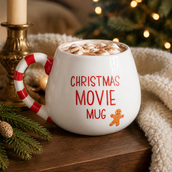 A white ceramic Christmas movie mug with a red and white candy cane handle, filled with a hot cocoa drink topped with marshmallows. The mug features the text 'CHRISTMAS MOVIE MUG' in red and a small gingerbread man graphic.