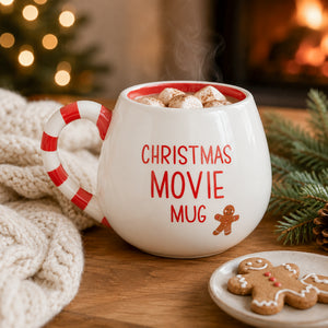 A white ceramic Christmas movie mug with a red and white candy cane handle, filled with marshmallows and hot cocoa. The mug features the text 'CHRISTMAS MOVIE MU' in red, next to a gingerbread cookie on a small plate, set on a wooden table with a cozy fireplace and holiday decorations in the background.