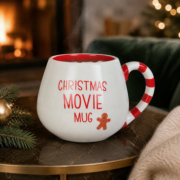 A white ceramic mug with a red interior and red text reading 'CHRISTMAS MOVIE MUG' plus a small gingerbread figure, shown on a table in a cozy holiday setting with a fireplace and green chair in the background.