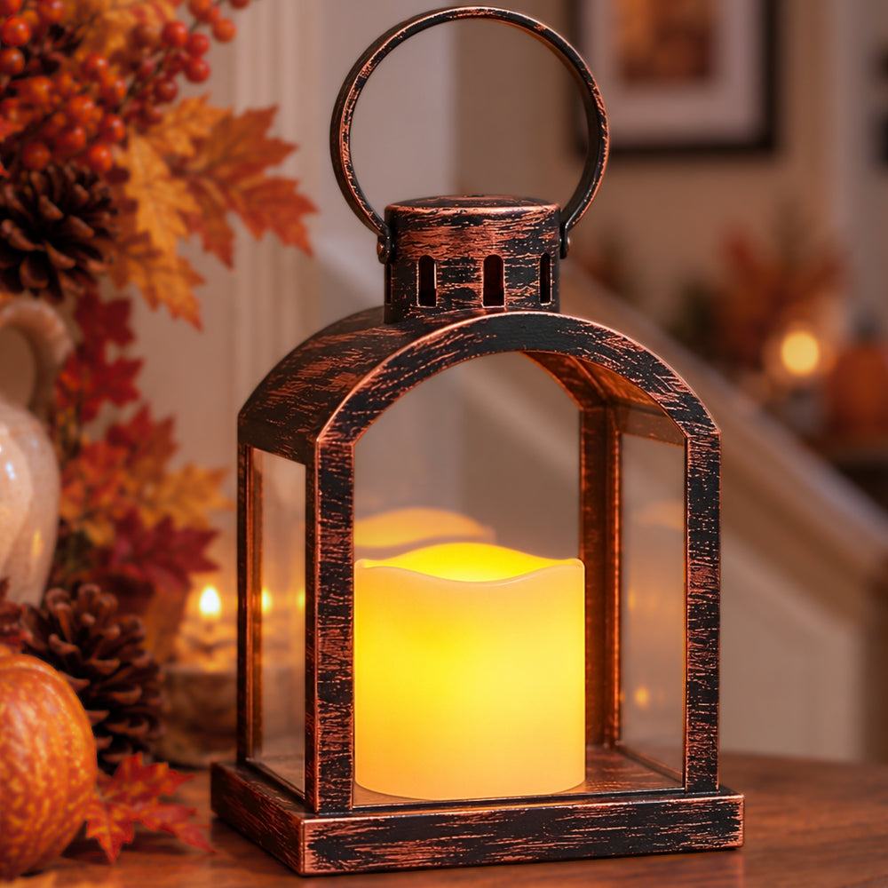 A rustic bronze lantern with a glowing yellow LED candle inside, set on a wooden table in a cozy, softly blurred home interior decorated with autumn leaves and pumpkins.