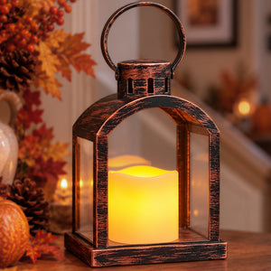 A rustic bronze lantern with a glowing yellow LED candle inside, set on a wooden table in a cozy, softly blurred home interior decorated with autumn leaves and pumpkins.