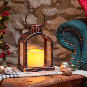 A rustic copper lantern with a glowing yellow LED candle inside, styled on a wooden table with Christmas ornaments, a stone wall background, and a teal velvet armchair.