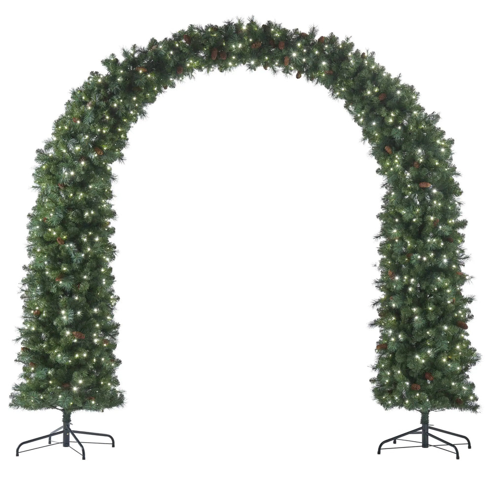 A festive holiday wreath with warm white string lights draped over a dark green double front door, set in a stone entryway with lit wall sconces and manicured hedges.