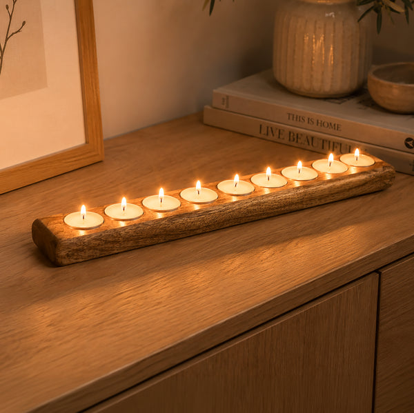 A rustic wooden candle holder with ten lit tealight candles, placed on a wooden cabinet in a warmly lit room with books and a framed picture in the background.