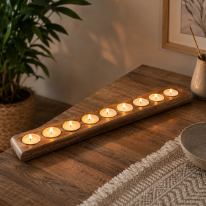 A long rectangular wooden candle holder with multiple small lit tealight candles, placed on a wooden table in a styled interior setting with a plant, bowl, and framed art visible.