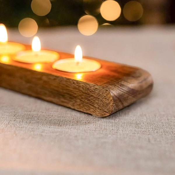 A close-up of a rectangular wooden candle holder with rounded corners, holding three lit tealight candles on a textured beige fabric surface with warm bokeh lights in the background.