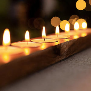 A rustic wooden tray holding several lit tealight candles, photographed in warm ambient light with soft bokeh background lights.