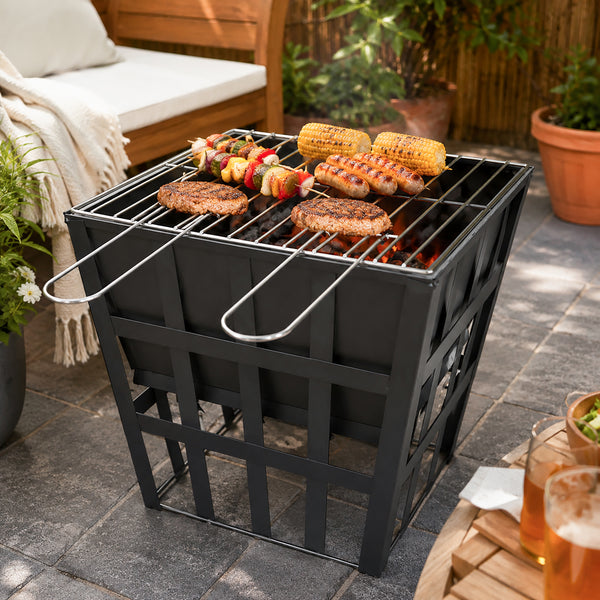 A black metal charcoal grill with a grate top, shown outdoors on a stone patio with food cooking on it. The grill has a slatted front and side panels, with metal handles extending from the sides.