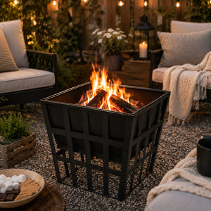 A black metal square fire pit with a lattice-style base, shown lit with orange flames and logs in an outdoor patio setting with chairs, pillows, candles, and string lights.