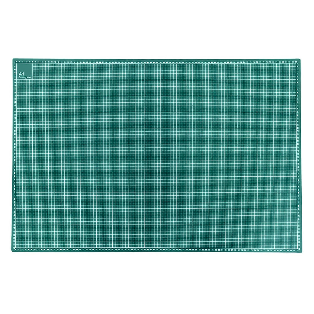 A person uses a metal ruler and pencil to measure and draw on a large green cutting mat on a wooden desk. The scene includes crafting supplies, a small plant, and a mint green machine in a bright, tidy workspace.