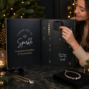 A woman opens a black advent calendar box labeled 'Twelve Days of Sparkle Gemstone Jewellery & Accessories' on a dark marble surface, with holiday lights and pine branches in the background. Gold-toned jewelry pieces are arranged nearby, and the calendar interior shows numbered compartments with moon-phase graphics.