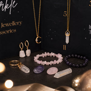 A styled flat-lay of jewelry accessories arranged on a dark surface with a branded black box behind them. Visible items include gold-tone necklaces, earrings, bracelets, a keychain, and several crystal stones in pink, purple, and clear tones.