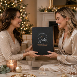 Two women in a warmly lit holiday living room smile while holding a black personalized jewelry box. The left woman wears a champagne sequined dress, and the right woman wears a champagne satin robe.