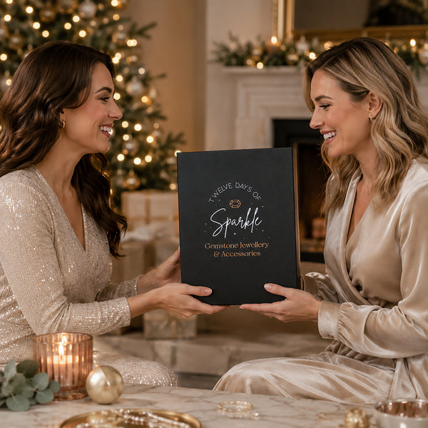 Two women in a warmly lit holiday living room smile while holding a black personalized jewelry box. The left woman wears a champagne sequined dress, and the right woman wears a champagne satin robe.