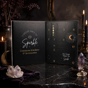 An open black advent calendar box labeled 'Twelve Days of Sparkle' with gold moon-phase and numbered door designs, displayed on a dark marble surface with candles, crystals, and dried flowers in the background.