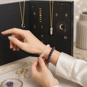 A person's hands are shown wearing two beaded bracelets: a light pink stone bracelet and a dark purple stone bracelet, in front of a black advent calendar displaying gold-toned moon-themed necklaces.