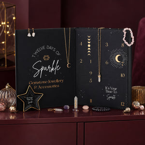 An open black advent calendar styled as a gemstone jewellery gift box, with gold moon phase graphics, numbered doors, hanging necklaces, earrings, crystals, and decorative props on a dark wooden surface against a deep burgundy background.
