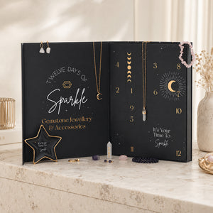 A black gemstone jewelry advent calendar styled on a marble countertop with crystals, gold-toned jewelry hanging from the flaps, and a neutral indoor background.