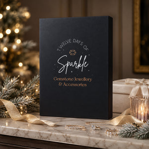 A black gift box with white and gold text reading 'Twelve Days of Sparkle Gemstone Jewellery & Accessories' sits on a marble surface in a warm holiday setting with a blurred Christmas tree, lights, ribbon, and wrapped gift box in the background.