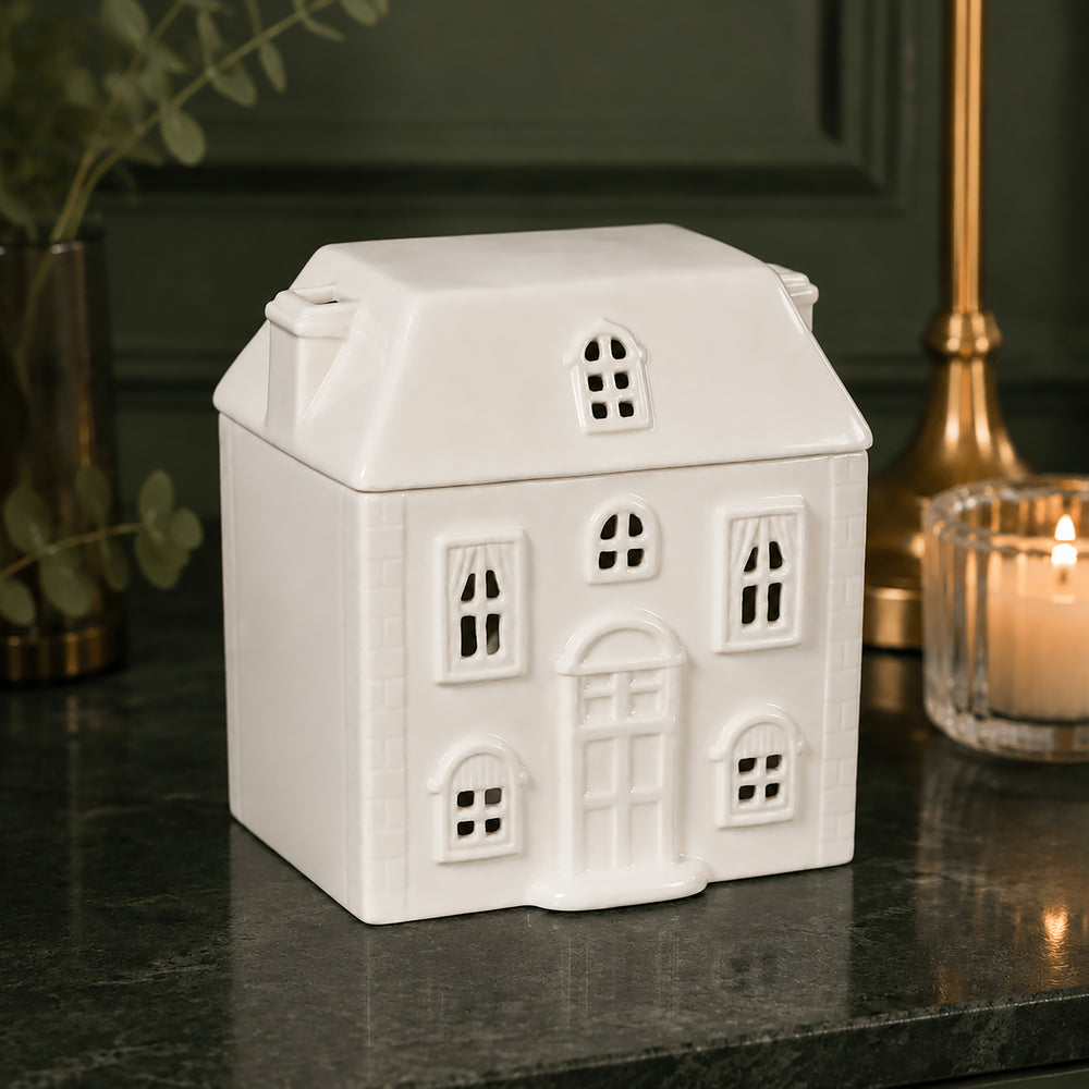 A white ceramic house-shaped tealight holder oil burner with cutout windows and door details, displayed on a dark marble surface next to a lit candle and greenery.