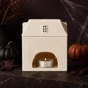 A cream-colored ceramic house-shaped candle holder with a lit silver tea light candle inside, styled on a dark stone surface with autumn pumpkins, spiderwebs, and dried leaves in the background.