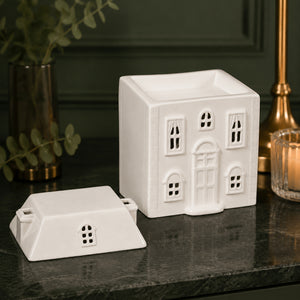 A white ceramic house-shaped candle holder with its matching lid placed beside it on a dark marble surface. The background features a dark green paneled wall, a gold candlestick, and a vase with greenery.