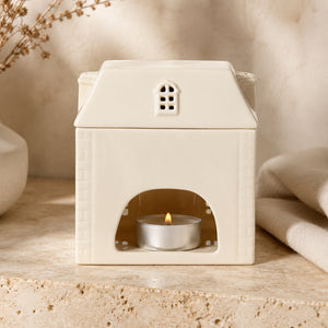 A cream-colored ceramic house-shaped candle warmer with a lit silver tea light candle inside, set on a textured stone surface with a soft beige background.