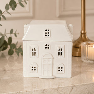 A small white ceramic house-shaped container or candle holder with cutout windows and a front door, displayed on a light stone surface with a candle and greenery in the background.