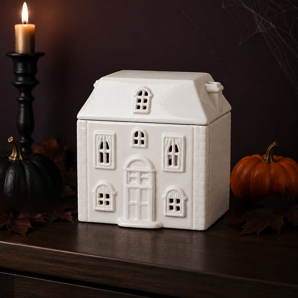 A white ceramic house-shaped cookie jar or decorative container with embossed window and door details, displayed on a dark wooden surface with a lit candle, pumpkins, and autumn leaves in the background.