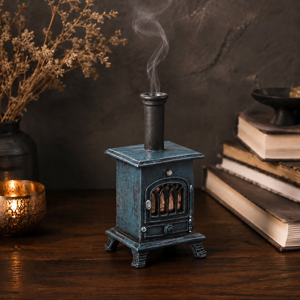 A miniature decorative wood-burning stove figurine in a distressed teal green finish, styled on a wooden table next to a  stack of books, candle and vase with dried flowers
