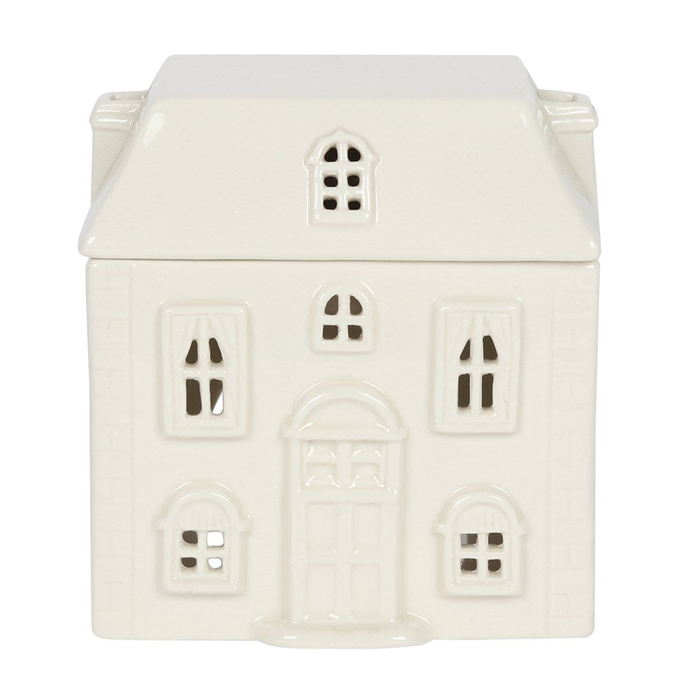 A white ceramic house-shaped tealight holder oil burner with cutout windows and door details, displayed on a dark marble surface next to a lit candle and greenery.