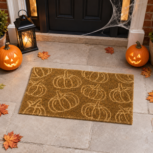 A rectangular coir doormat with a repeating pumpkin pattern, displayed on a stone patio in front of a black door. The scene is decorated for Halloween with two glowing jack-o'-lanterns, a black lantern, spiderwebs, and scattered autumn leaves.