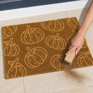 A rectangular coir doormat with a tan pumpkin pattern, shown on a light tile floor with a hand using a wooden brush to sweep dirt off the front.