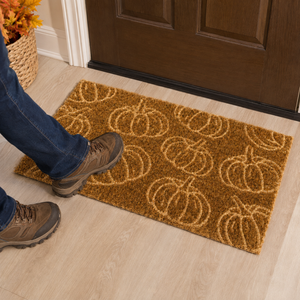 A rectangular brown coir doormat with a repeating pumpkin pattern, shown on a light wood floor in front of a dark brown door. A person's legs and feet wearing blue jeans and brown hiking boots are stepping onto the mat.