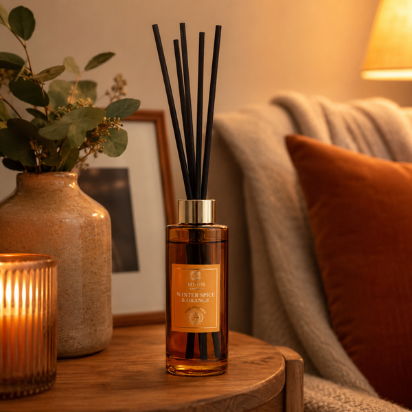 A reed diffuser bottle with black reeds and a warm amber liquid sits on a wooden side table in a cozy living room setting, with a lit candle, vase with greenery, framed art, and a sofa in the background.
