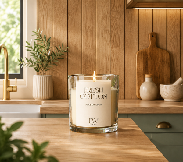 A lit scented candle in a clear glass jar sits on a wooden kitchen counter, with a wood-paneled wall, plants, and kitchen accessories in the background. The candle label reads 'FRESH COTTON' and 'Flour de Coton' with 'EW' at the bottom.