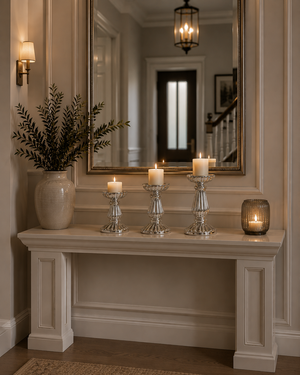 A styled entryway console table scene with a large ornate mirror, silver candlesticks with lit white candles, a small amber glass candle holder, a ceramic vase with dried greenery, and warm wall lighting.