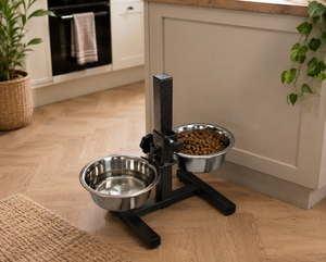 A black automatic pet feeder with two stainless steel bowls on a wooden kitchen floor, with one bowl filled with dry dog food and the other empty water bowl. The background shows a modern kitchen with light wood floors and cream-colored cabinets.