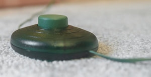 A close-up shot of a dark green, circular electronic device with a matching green button on top, resting on a textured white carpet with a thin wire extending from the side.