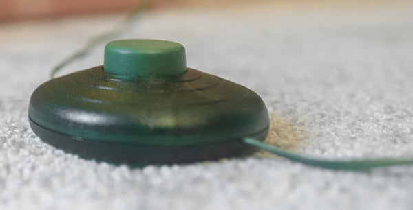 A close-up shot of a dark green, circular electronic device with a matching green button on top, resting on a textured white carpet with a thin wire extending from the side.