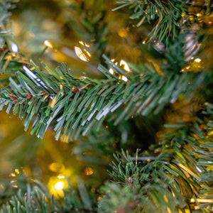 Close-up of an artificial Christmas tree branch with green needles and warm yellow string lights glowing in the background.