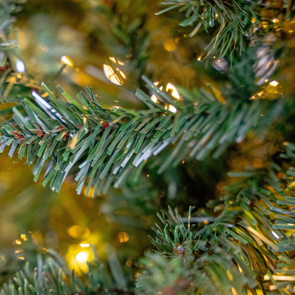 Close-up of an artificial Christmas tree branch with green needles and warm yellow string lights glowing in the background.