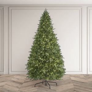 A tall, realistic artificial Christmas tree with dense green foliage and warm white string lights, standing on a black metal stand against an elegant white paneled wall and herringbone wood floor.