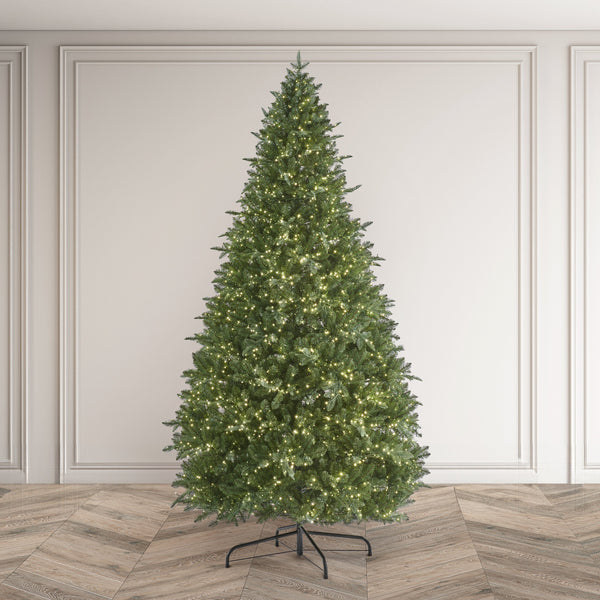 A tall, realistic artificial Christmas tree with dense green foliage and warm white string lights, standing on a black metal stand against an elegant white paneled wall and herringbone wood floor.