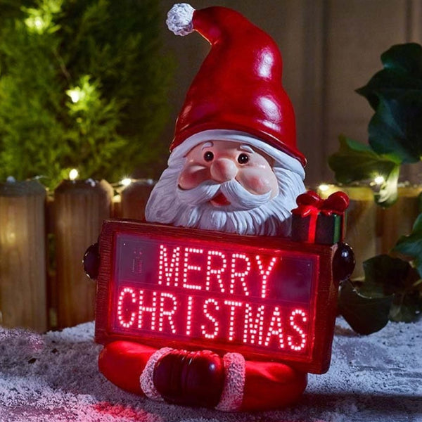 A festive Santa Claus garden ornament holding a glowing red LED sign that reads 'MERRY CHRISTMAS'. The ornament features a glossy red hat, white beard, and red suit, set against a softly blurred holiday background with warm lights and greenery.