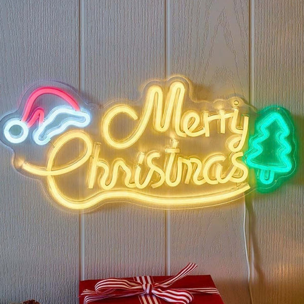 A glowing LED neon-style Christmas sign on a light wood-paneled wall, with a yellow 'Merry Christmas' script, a pink and white Santa hat on the left, a green Christmas tree on the right, and a red gift box with a striped bow at the bottom.