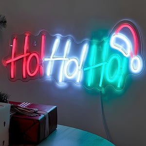 A glowing neon-style wall sign spelling "Holly Holly" in red, white, and teal, with a white neon Santa hat at the end, mounted on a wall above a table with wrapped gifts.
