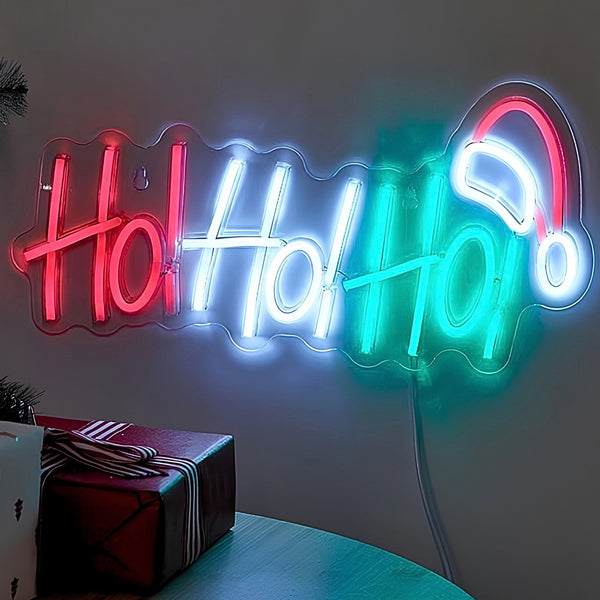 A glowing neon-style wall sign spelling "Holly Holly" in red, white, and teal, with a white neon Santa hat at the end, mounted on a wall above a table with wrapped gifts.