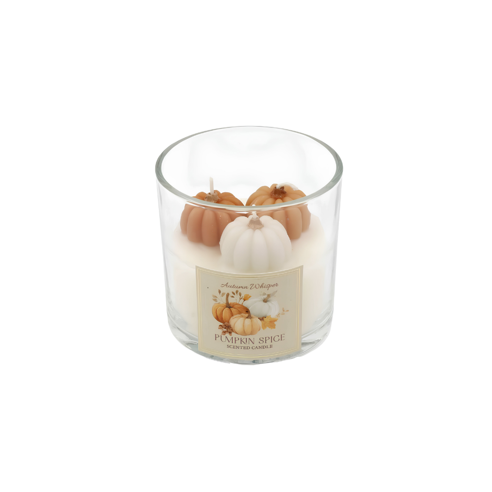 A glass jar candle labeled 'Autumn Village Pumpkin Spice Longburn Candle' sits on a round beige tray on a wooden table in a cozy living room setting. The candle features cream-colored wax and decorative pumpkins in orange, white, and light green on top.