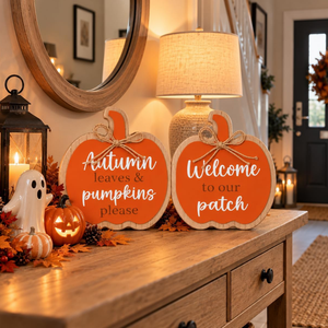 Two orange autumn pumpkin-shaped signs with white script text reading 'Autumn leaves & pumpkins please' and 'Welcome to our patch,' displayed on a wooden console table in a warmly lit entryway with a lamp, lantern, ghost decoration, and fall decor.